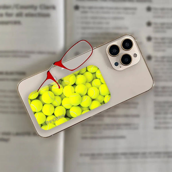 Red ThinOptics reading glasses rest atop a smartphone case displaying a vibrant pattern of bright yellow tennis balls.