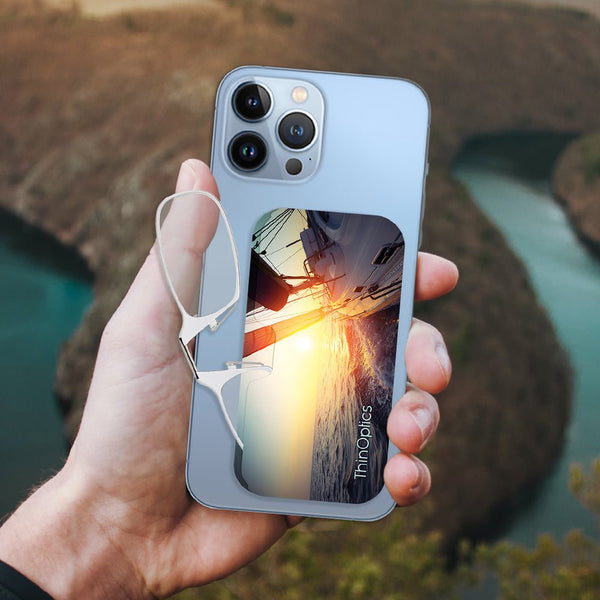 Hand holds a blue smartphone with ThinOptics reading glasses attached. The phone displays a vibrant sunset image of sailboats racing on the water.