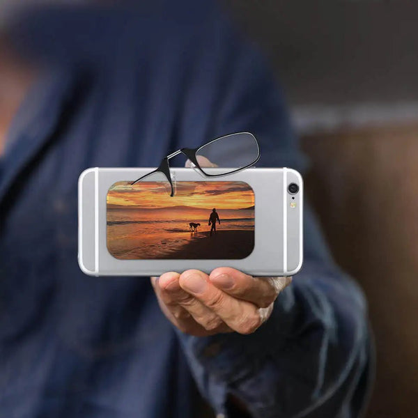 This image displays a pair of black-rimmed Readers alongside a glasses case. The glasses case features a scenic photograph depicting a person walking a dog along a beach during a vibrant sunset. The sky is filled with warm hues of orange, yellow, and pink, which reflect off the water, creating a serene and picturesque atmosphere. The silhouettes of the person and the dog add a sense of tranquility and companionship to the image.