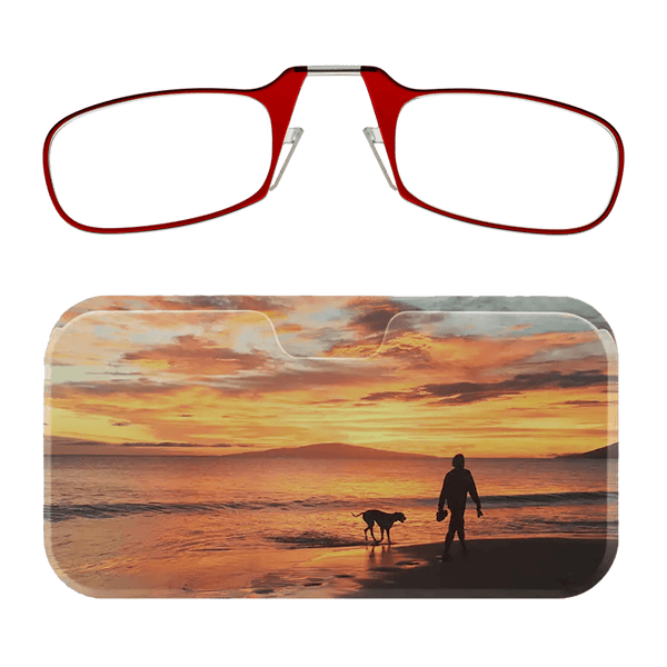 This image displays a pair of red-rimmed Readers alongside a glasses case. The glasses case features a scenic photograph depicting a person walking a dog along a beach during a vibrant sunset. The sky is filled with warm hues of orange, yellow, and pink, which reflect off the water, creating a serene and picturesque atmosphere. The silhouettes of the person and the dog add a sense of tranquility and companionship to the image.