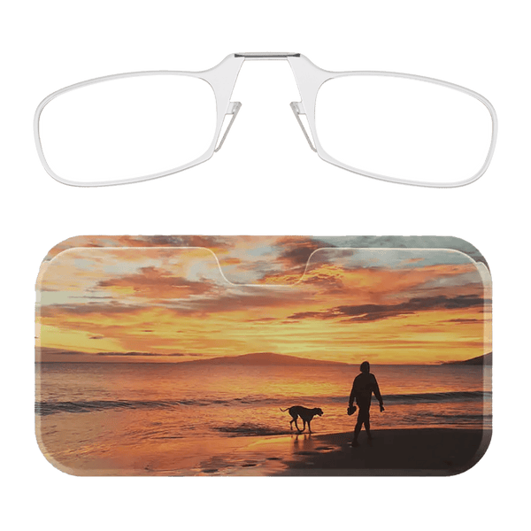 This image displays a pair of clear-rimmed Readers alongside a glasses case. The glasses case features a scenic photograph depicting a person walking a dog along a beach during a vibrant sunset. The sky is filled with warm hues of orange, yellow, and pink, which reflect off the water, creating a serene and picturesque atmosphere. The silhouettes of the person and the dog add a sense of tranquility and companionship to the image.