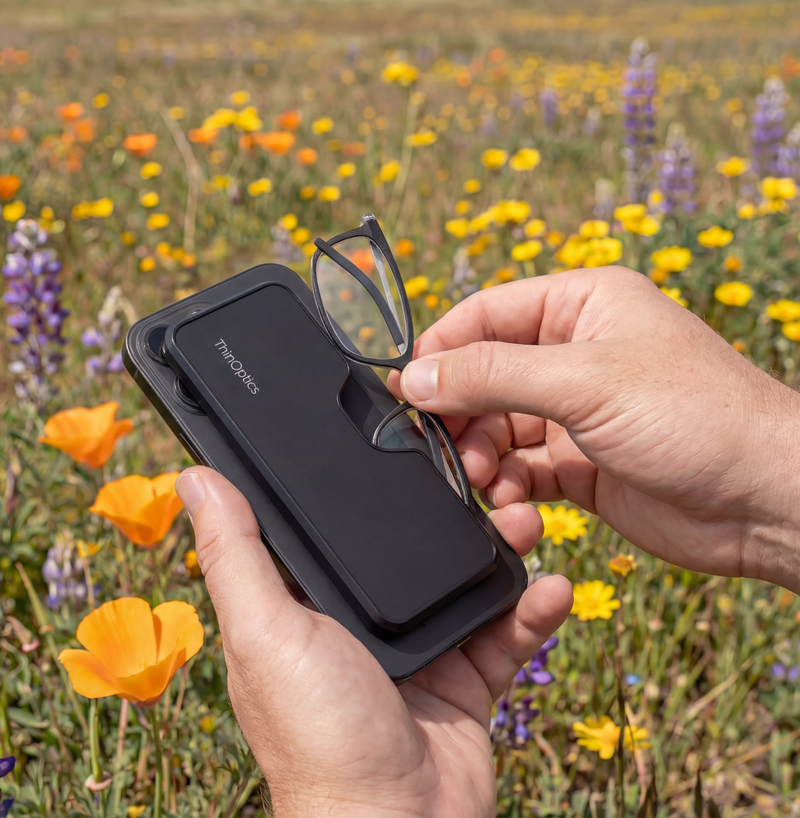Hand pulling ThinOptics readers from a MagSafe reading glasses case attached to a smartphone in a flower field.
