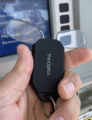 A hand holding a Readers + Keychain Case in front of a gas station.