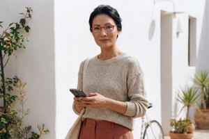 Woman wearing red ThinOptics reading glasses while looking at her smartphone in a sunlit outdoor courtyard.