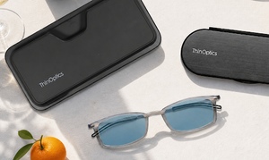 ThinOptics blue light blocking glasses beside a smartphone and slim carrying case on a sunlit wooden table.