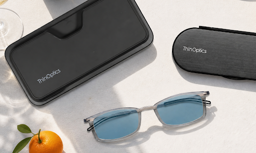 ThinOptics blue light blocking glasses beside a smartphone and slim carrying case on a sunlit wooden table.