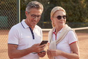 Couple wearing ThinOptics reading sunglasses while looking at a phone together on a sunny outdoor tennis court.