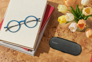 Blue ThinOptics glasses rest on stacked books beside a black case and a vase of tulips.
