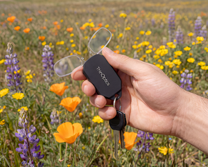 Hand holding ThinOptics reading glasses in a keychain case attached to keys in a garden setting.