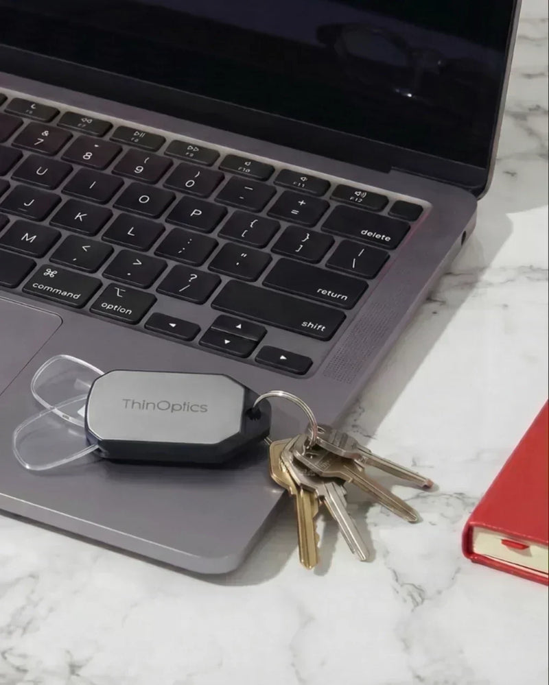 ThinOptics keychain case with compact reading glasses resting beside a laptop and keys on a workspace, ready for everyday screen use.