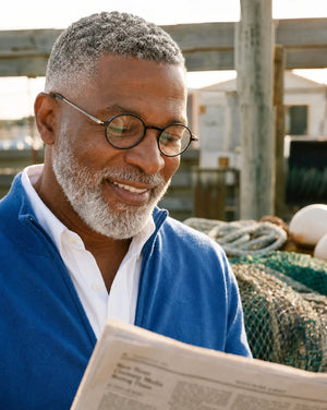 Man wearing ThinOptics prescription glasses smiles while reading a newspaper outdoors near a marina, demonstrating comfortable vision for everyday activities.