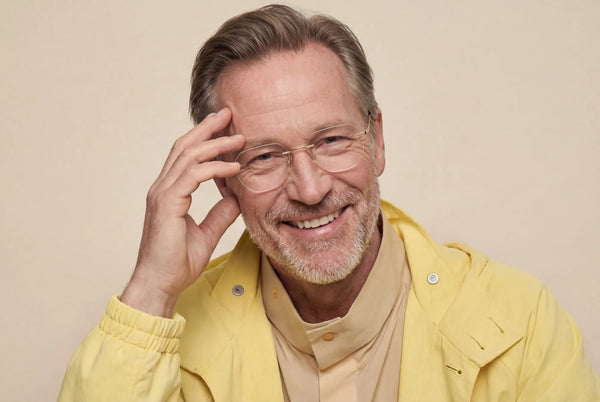 Smiling bearded man in a yellow jacket adjusting his rimless gold-frame reading glasses with both hands against a neutral studio background