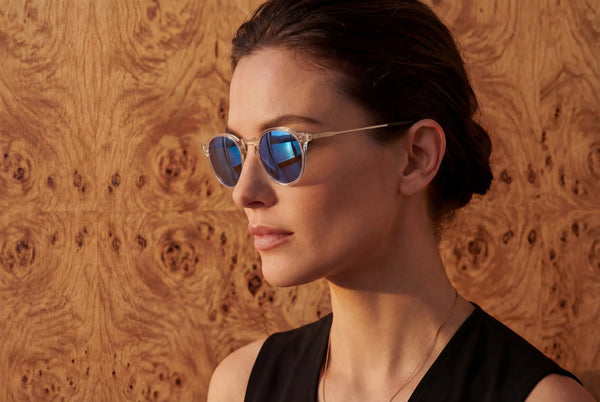 Close-up profile of a woman wearing round sunglasses with clear frames and blue mirrored lenses, against a warm burl wood background