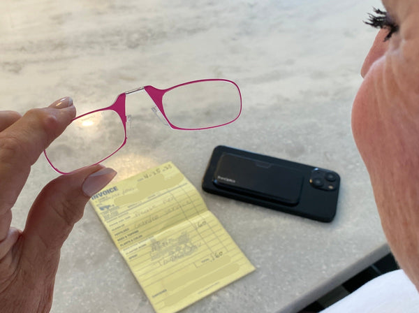 Woman holding Blushing Pink Spring Bloom Readers at marble kitchen island, reviewing invoice, with Black Universal Pod Case nearby - Limited Edition ThinOptics Reading Glasses