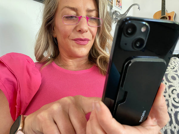 Woman wearing Blushing Pink Readers while reading her iPhone with Black Universal Pod on back