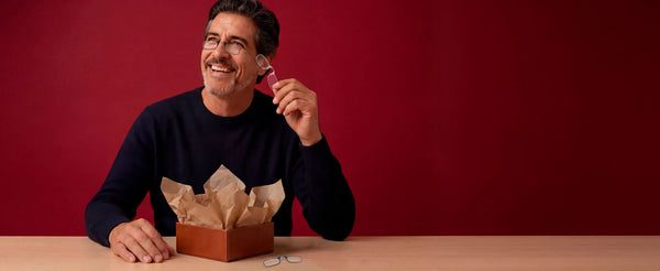 Smiling man seated at table against red background holding compact folding reading glasses from gift box