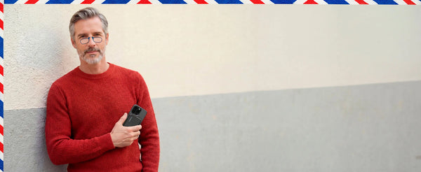 Man in red sweater wearing ThinOptics reading glasses holds smartphone with attached ThinOptics reader case while standing against neutral wall