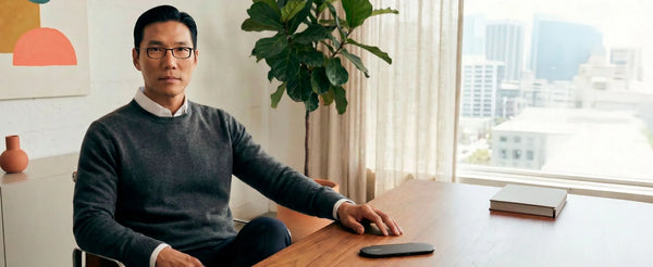 Man wearing ThinOptics reading glasses seated at desk near window with ThinOptics slim reader case placed on wooden table