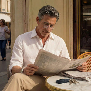 Man in a white linen shirt wearing armless reading glasses reading a newspaper at a sunlit Paris cafe table with a ThinOptics keychain case on his keys