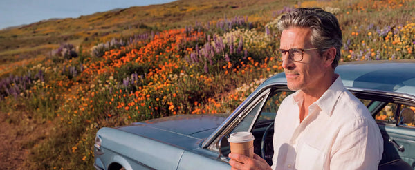 Man wearing tortoiseshell reading glasses leaning on a vintage car, holding coffee, with a colorful wildflower hillside behind him