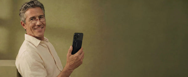 Smiling man wearing red reading glasses holding a smartphone with a ThinOptics phone case attached to the back, against a warm olive background