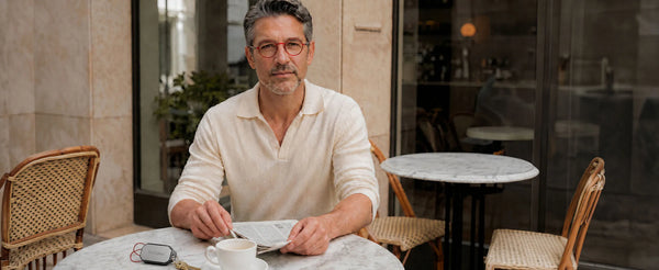 Man in a cream sweater wearing red round reading glasses at a marble cafe table with a newspaper, coffee, and ThinOptics keychain case on his keys