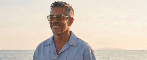 Smiling silver-haired man wearing tortoiseshell aviator sunglasses with gold frames, standing by the ocean at golden hour in a denim shirt