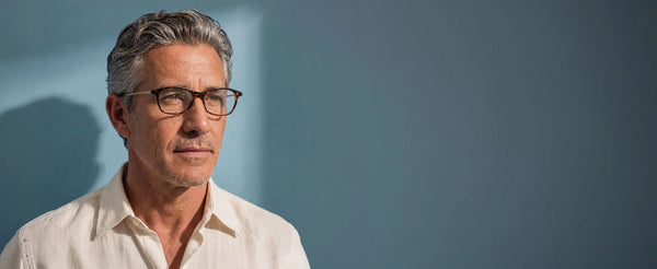 Silver-haired man in a linen shirt wearing tortoiseshell rectangular prescription glasses gazing to the side against a soft blue wall with shadow detail