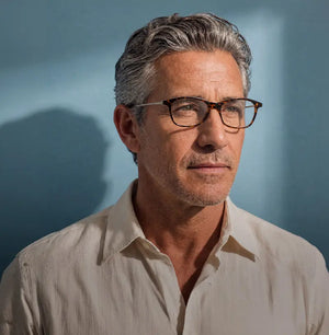 Silver-haired man in a linen shirt wearing tortoiseshell rectangular prescription glasses gazing to the side against a soft blue wall with shadow detail