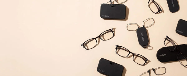 ThinOptics black reading glasses collection flatlay on cream background featuring multiple frame styles with pod cases and keychain cases scattered