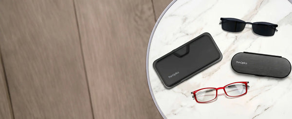 ThinOptics Brooklyn red reading glasses, black sunglasses, black Pod case, and Brooklyn case arranged on a marble side table