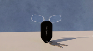 ThinOptics clear armless reading glasses peeking out of a black keychain case with keys attached, against a blue backdrop