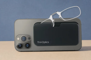 ThinOptics clear armless reading glasses peeking out of a black MagSafe phone case on the back of an iPhone, side view