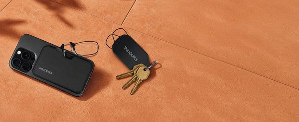 ThinOptics Complete Carry Bundle flatlay showing black MagSafe phone case with readers and black keychain case with keys on an orange surface