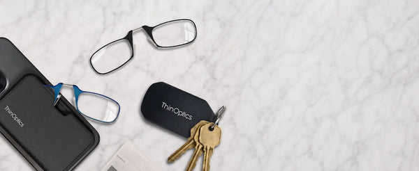 ThinOptics flatlay of black and blue armless readers, black MagSafe phone case, and black keychain case with keys on white marble