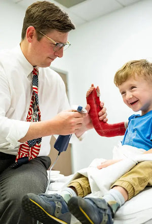Doctor wearing ThinOptics clear glasses removing a red cast from a smiling young boy's arm in a medical office