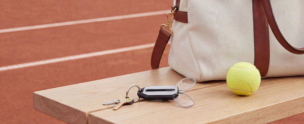 ThinOptics keychain case with compact reading glasses attached to keys on a wooden bench, beside a tennis ball and canvas bag on a clay court
