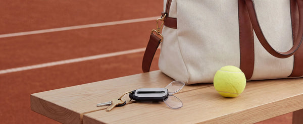 ThinOptics keychain case with compact reading glasses attached to keys on a wooden bench, beside a tennis ball and canvas bag on a clay court