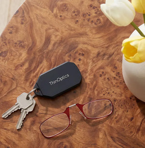 ThinOptics keychain case attached to keys beside red armless reading glasses on a burl wood table, with a vase of colorful tulips nearby