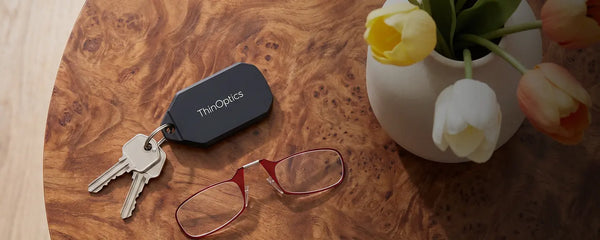 ThinOptics keychain case attached to keys beside red armless reading glasses on a burl wood table, with a vase of colorful tulips nearby