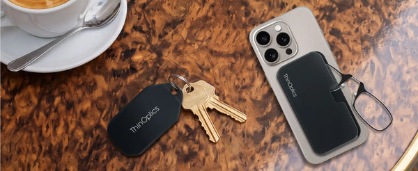 ThinOptics black keychain case with keys and MagSafe phone case with armless readers on a burlwood cafe table beside a coffee cup