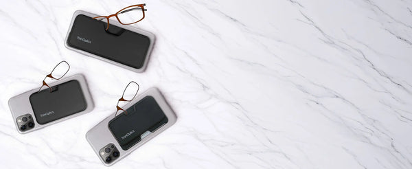 Three iPhones with ThinOptics magnetic MagSafe cases and brown reading glasses peeking out, arranged on a white marble surface