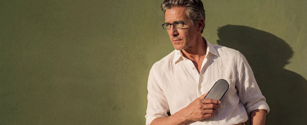 Man wearing ThinOptics black rectangle glasses holding a ThinOptics Brooklyn case against a sun-lit green stucco wall