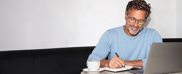 Smiling man wearing ThinOptics black rectangle reading glasses writing in a notebook at a café table with a laptop and coffee cup
