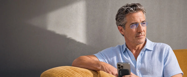Man wearing ThinOptics Moment blue light blocking readers holding an iPhone with a ThinOptics Pod case on a yellow sofa