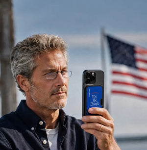 Man wearing ThinOptics reading glasses holding an iPhone with a ThinOptics We the People MagSafe case, American flag in the background