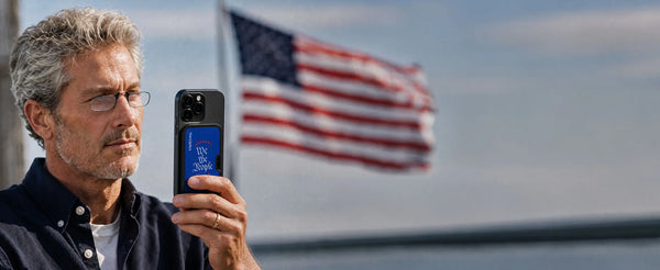 Man wearing ThinOptics reading glasses holding an iPhone with a ThinOptics We the People MagSafe case, American flag in the background