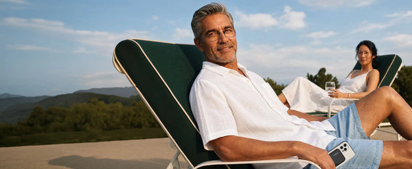 Man wearing ThinOptics glasses relaxing on a lounge chair outdoors with a ThinOptics phone case, woman with a drink in the background