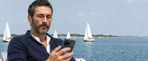 Man wearing ThinOptics reading glasses using his phone with a blue Americana-print MagSafe case, sailboats on the water behind him