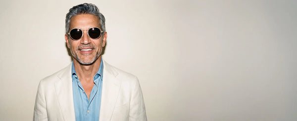Smiling man wearing ThinOptics gold round sunglasses and a white blazer against a neutral background